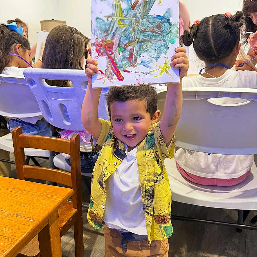 Enzo Toro is registered to the contest to win money with this photo: art_class, boy, chairs, child, colorful, creative, drawing, expression, floor, hair, happy, indoor, kids, painting, people, playful, shirt, shorts, smiling, wooden_table
