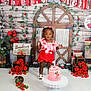 toddler, child, birthday_cake, cake_stand, red_roses, strawberries, flower_garland, decorations, party, celebration, pink_dress, white_sandals, wooden_background, green_leaves, sign, flowers, standing, indoors, happy, festive