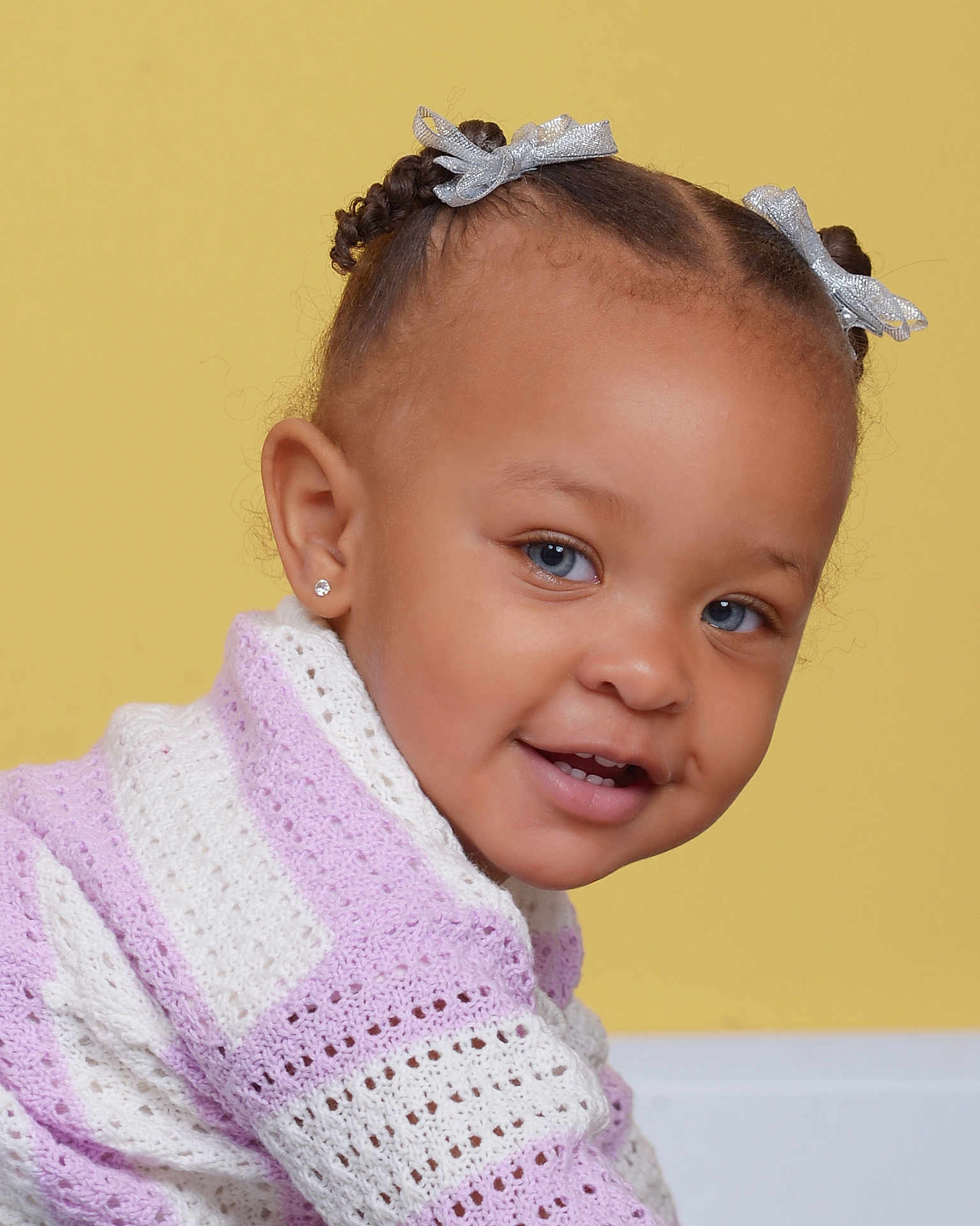 Bria is registered to the contest to win money with this photo: toddler, child, smile, face, blue_eyes, braided_hair, silver_bows, purple_sweater, white_sweater, knitwear, earrings, portrait, cute, happy, indoor, yellow_background, baby, fashion, closeup, person