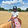arena, braids, child, clouds, colorful_dress, daylight, equestrian, fence, happy, helmet, horse, nature, outdoor, person, pony, riding, sandy_ground, sky, smiling, trees