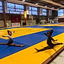child, gymnast, gymnasium, sports, floor_mat, yellow_mat, blue_mat, split_pose, flexibility, young_girl, athlete, indoor, sportswear, medal, competition, banners, trophies, wooden_beams, lighting, training