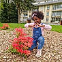 child, daylight, denim_overalls, flowers, garden, girl, grass, greenery, heart_shape, nature, outdoor, pink_headband, playful, portrait, red_bush, residential_building, smiling, sneakers, tree, wood_chips