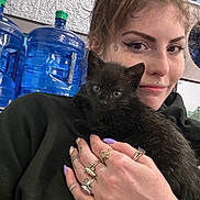 Wolfie is registered to the contest to win money with this photo: person, cat, black_cat, kitten, hand, rings, tattoo, nail_polish, water_jug, container, wall, indoor, casual, portrait, face, animal, pet, holding, closeup, smile