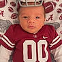 Raylan is registered to the contest to win money with this photo: baby, infant, football_helmet, football_jersey, alabama, blanket, elephant_pattern, sportswear, cute, portrait, lying_down, child, red, white, face, headgear, apparel, smiling, young_child, cozy
