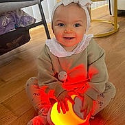 Loudjayne participe au concours pour gagner de l'argent avec cette photo : baby, child, glowing_lamp, headband, bow, smiling, sitting, floor, wooden_floor, leggings, patterned_clothing, cozy, indoors, table, remote_control, furniture, happy, cute, hands, face