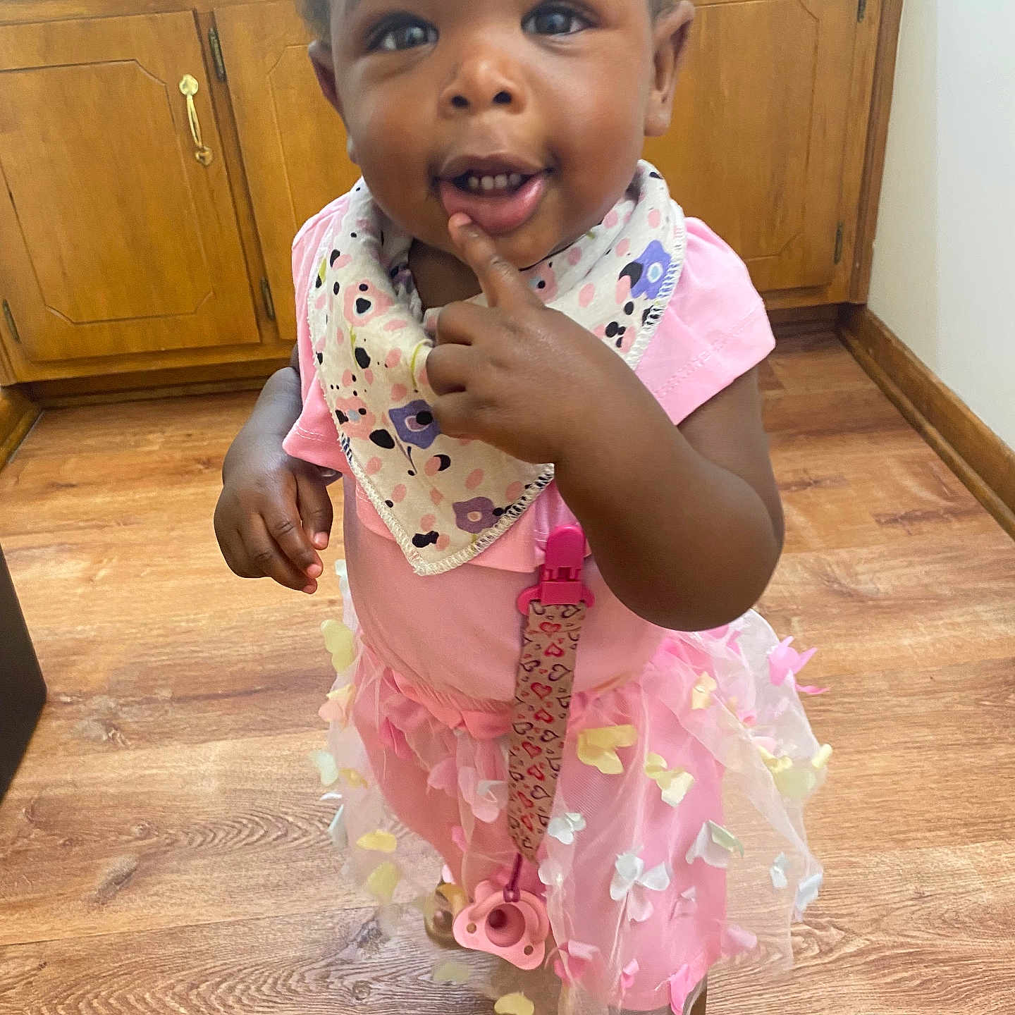 Emersyn joined the competition — help win amazing prizes! toddler, child, pink_dress, tutu, bib, floor, wooden_floor, wooden_cabinets, indoors, curious, standing, pacifier, clip, finger_on_lip, cute, smiling, shoes, person, baby, home