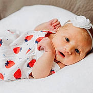 Brooklynn joined the competition — help win amazing prizes! baby, infant, newborn, white_dress, strawberry_pattern, headband, flower, soft_cushion, lying_down, curious_expression, cute, close_up, indoor, skin, small_hands, face, eyes, mouth, resting, comfortable