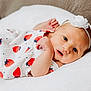 baby, infant, newborn, white_dress, strawberry_pattern, headband, flower, soft_cushion, lying_down, curious_expression, cute, close_up, indoor, skin, small_hands, face, eyes, mouth, resting, comfortable