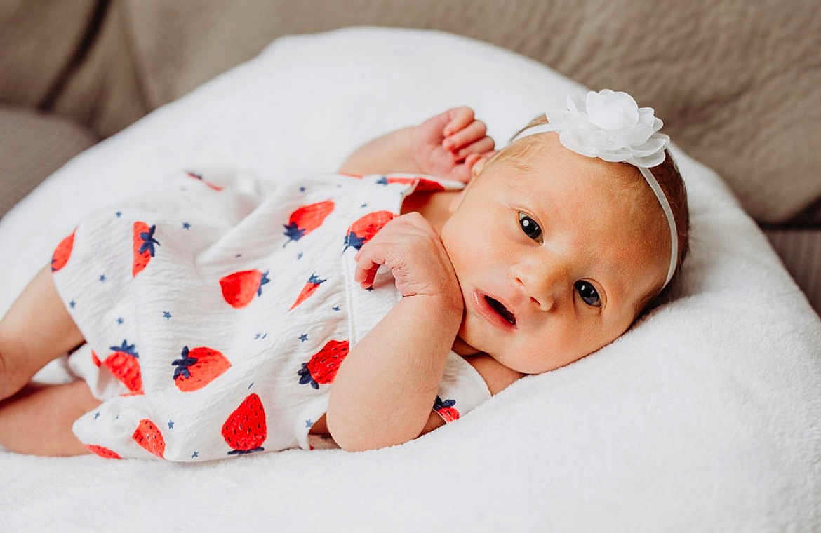 Brooklynn joined the competition — help win amazing prizes! baby, infant, newborn, white_dress, strawberry_pattern, headband, flower, soft_cushion, lying_down, curious_expression, cute, close_up, indoor, skin, small_hands, face, eyes, mouth, resting, comfortable