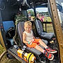 backpack, boy, casual_clothing, child, daytime, grass, helicopter, helmet, military, orange_shorts, outdoor, people, pilot_seat, red_sneakers, seat, sky, smile, sunny, vehicle, window