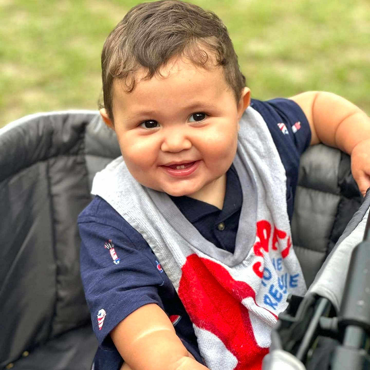 Raylan is registered to the contest to win money with this photo: arm, baby, bib, casual, child, clothing, cute, face, grass, hand, happy, infant, outdoor, person, portrait, seat, short_hair, skin, smiling, stroller