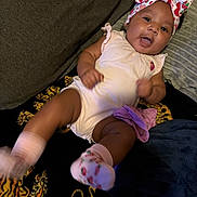 Nalani joined the competition — help win amazing prizes! baby, infant, child, portrait, smiling, curious, sitting, lying_down, indoors, couch, blanket, headband, bow, floral_pattern, strawberry, romper, socks, dark_background, home, face