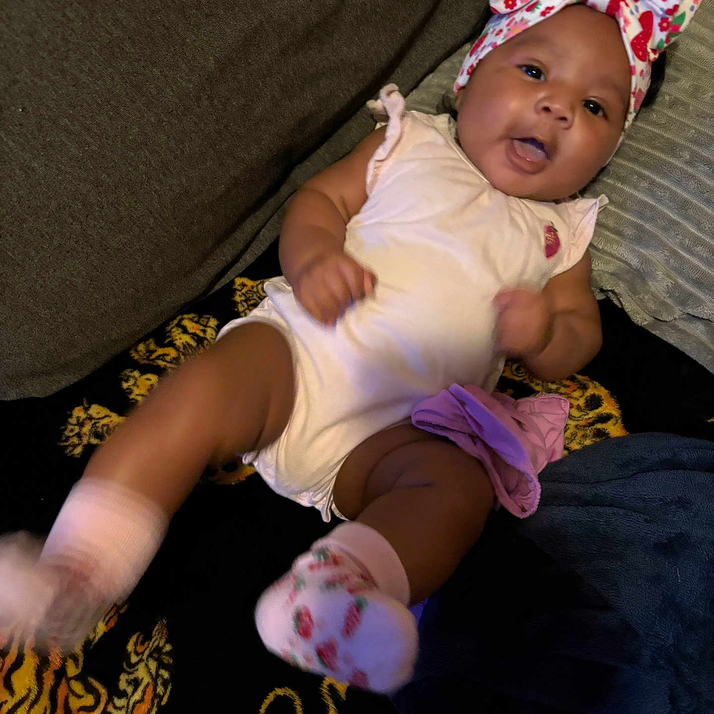 Nalani joined the competition — help win amazing prizes! baby, infant, child, portrait, smiling, curious, sitting, lying_down, indoors, couch, blanket, headband, bow, floral_pattern, strawberry, romper, socks, dark_background, home, face