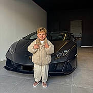 émilio participe au concours pour gagner de l'argent avec cette photo : child, boy, smile, sports_car, lamborghini, garage, puffy_jacket, blond_hair, standing, posing, headlight, luxury, concrete_floor, door, hood, front_view, casual_shoes, portrait, indoor, happy