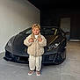 child, boy, smile, sports_car, lamborghini, garage, puffy_jacket, blond_hair, standing, posing, headlight, luxury, concrete_floor, door, hood, front_view, casual_shoes, portrait, indoor, happy