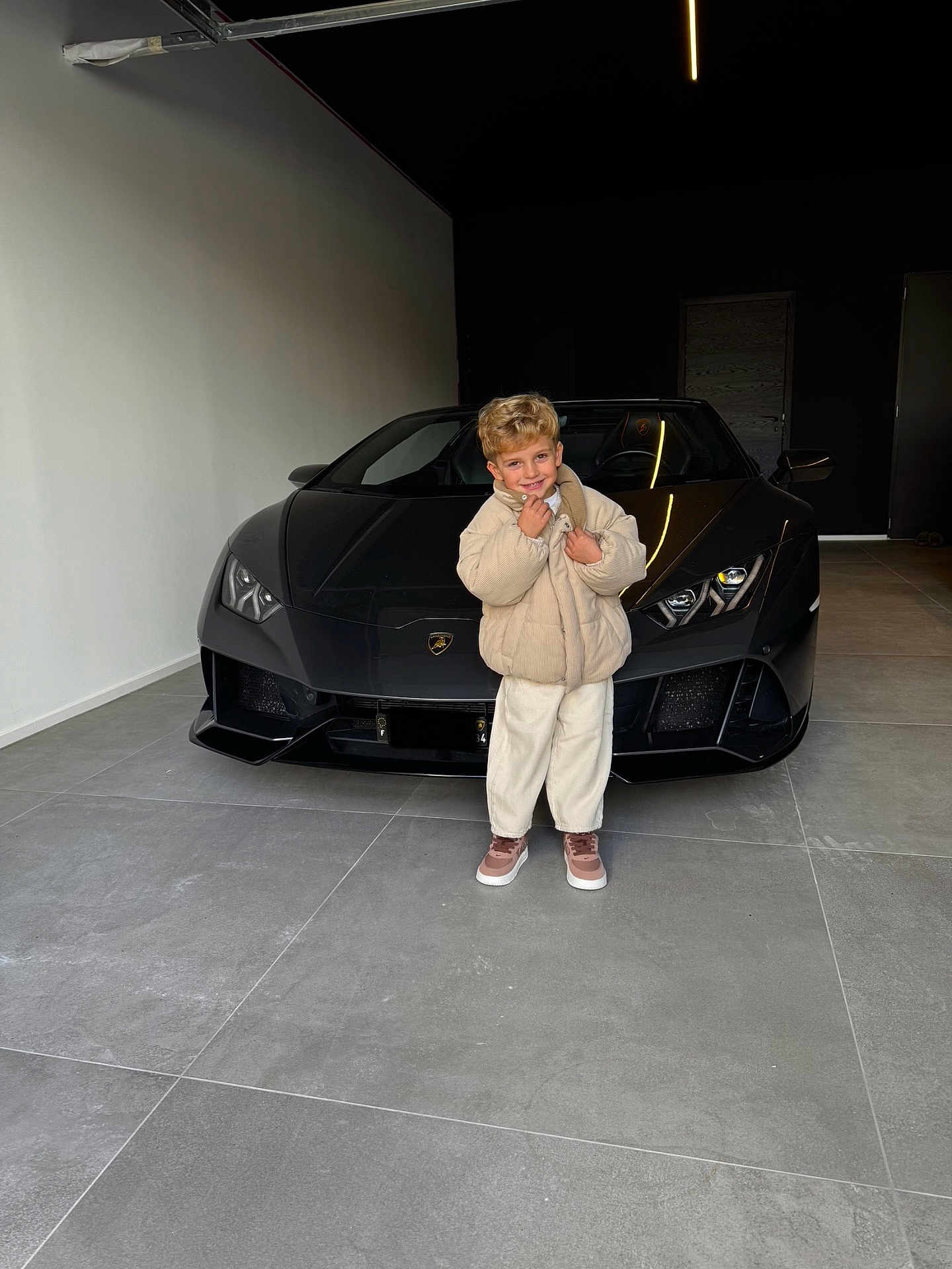 émilio participe au concours pour gagner de l'argent avec cette photo : child, boy, kid, car, sports_car, lamborghini, luxury, garage, concrete_floor, coat, smile, portrait, standing, shoes, head, blonde_hair, vehicle, headlights, tiled_floor, black_car
