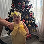 baby, child, yellow_dress, headband, socks, christmas_tree, ornaments, indoor, curtains, floor, hand, support, holiday, decorations, window, light, person, standing, festive, home