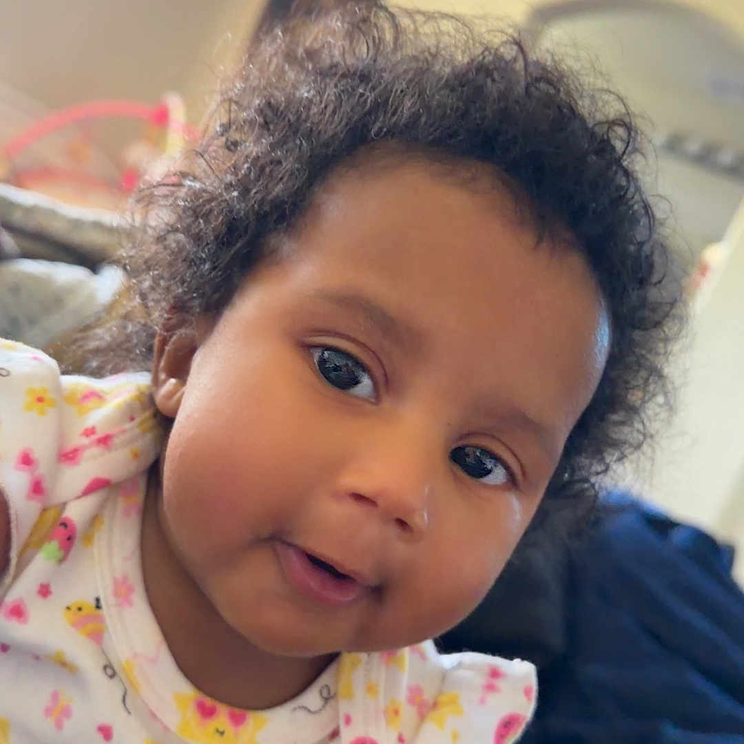 Stacia is registered to the contest to win money with this photo: baby, child, curly_hair, onesie, patterned_clothing, indoor, blanket, bedroom, face, hand, infant, portrait, cute, close_up, person, toddler, soft_light, smiling, young_child, resting