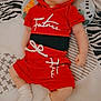 Christopher is registered to the contest to win money with this photo: baby, child, red_clothing, hoodie, shorts, white_socks, lying_down, pillow, bedspread, patterned_bed, infant, cute, sleepy, indoors, young_child, resting, casual_wear, portrait, small_person, relaxed