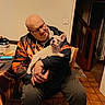man, glasses, hoodie, dog, puppy, french_bulldog, indoor, chair, table, tile_floor, wooden_furniture, warm_lighting, person, pet, sitting, holding, smiling, casual_clothing, home, cozy