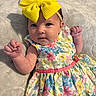 baby, child, headband, yellow_bow, dress, floral_pattern, colorful_clothing, blanket, fist, cute, infant, portrait, soft_texture, laying_down, indoors, person, skin, face, expression, small