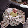 baby, blanket, couch, floral_pattern, indoors, infant, person, cozy, furniture, carpet, floor, slippers, plant, home, relaxing, yellow_clothing, cute, wrapped, resting, soft