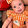 Kori joined the competition — help win amazing prizes! baby, infant, child, portrait, smiling, tongue_out, blue_eyes, bow, checkered_outfit, orange_clothing, romper, sitting, close_up, indoors, cushion, adult_arm, happy, cute, fashion, photography