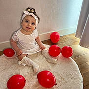 Sofia participe au concours pour gagner de l'argent avec cette photo : baby, child, girl, white_dress, headband, red_balloons, sitting, rug, wooden_floor, curtains, indoor, smiling, looking_up, cute, happy, decor, soft_light, portrait, floor, cozy