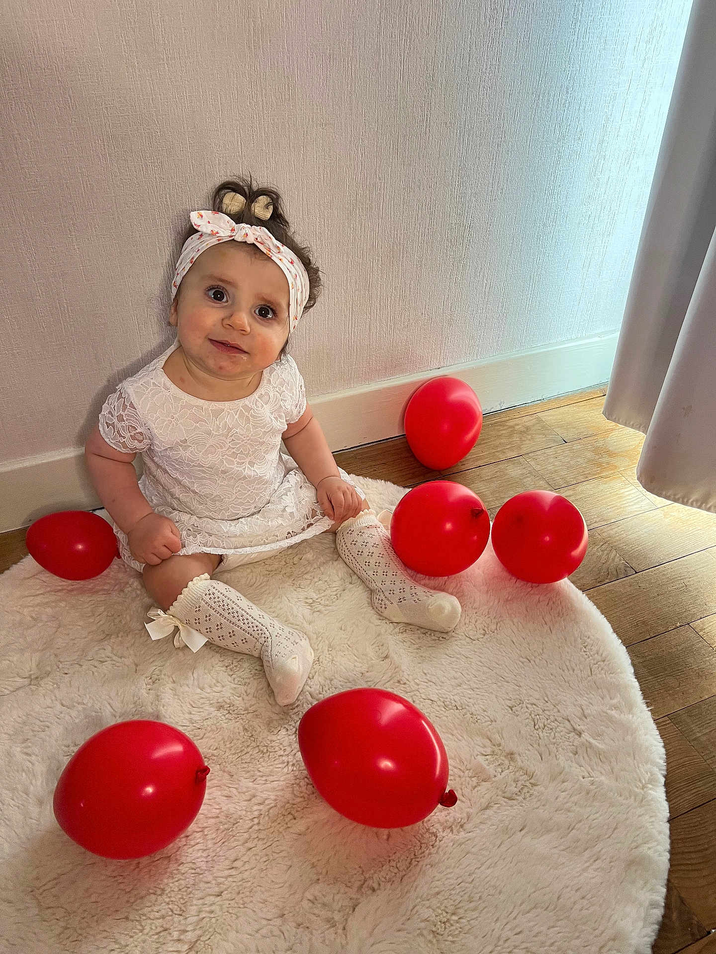 Sofia participe au concours pour gagner de l'argent avec cette photo : baby, child, girl, white_dress, headband, red_balloons, sitting, rug, wooden_floor, curtains, indoor, smiling, looking_up, cute, happy, decor, soft_light, portrait, floor, cozy