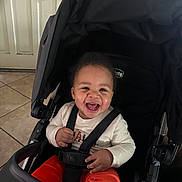 King is registered to the contest to win money with this photo: baby, baby_face, black_stroller, child, clothing, cute, door, happy, indoor, infant, joyful, orange_pants, person, portrait, seatbelt, sitting, smiling, stroller, tiled_floor, white_shirt