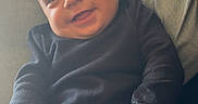 Kruz is registered to the contest to win money with this photo: baby, child, infant, smiling, portrait, sitting, sofa, couch, indoors, home, sunlight, window, black_clothing, dark_outfit, hands, barefoot, face, head, happy, cozy