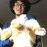 person, cat, indoor, curly_hair, glasses, smiling, holding, orange_cat, white_cat, fluffy_cat, playful, funny_pose, ceiling_light, casual_clothing, home, portrait, happy, pet, cute, animal