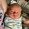 River is registered to the contest to win money with this photo: baby, newborn, infant, swaddle, tattoo, adult_arm, indoor, person, face, expression, curious, blanket, furniture, table, clothing, pattern, skin, head, holding, closeup