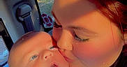 River is registered to the contest to win money with this photo: baby, woman, kiss, car_interior, blue_onesie, sunlight, face, child, portrait, love, affection, seatbelt, window, nature_outside, casual_clothing, closeup, parent_and_child, human, indoor, happy
