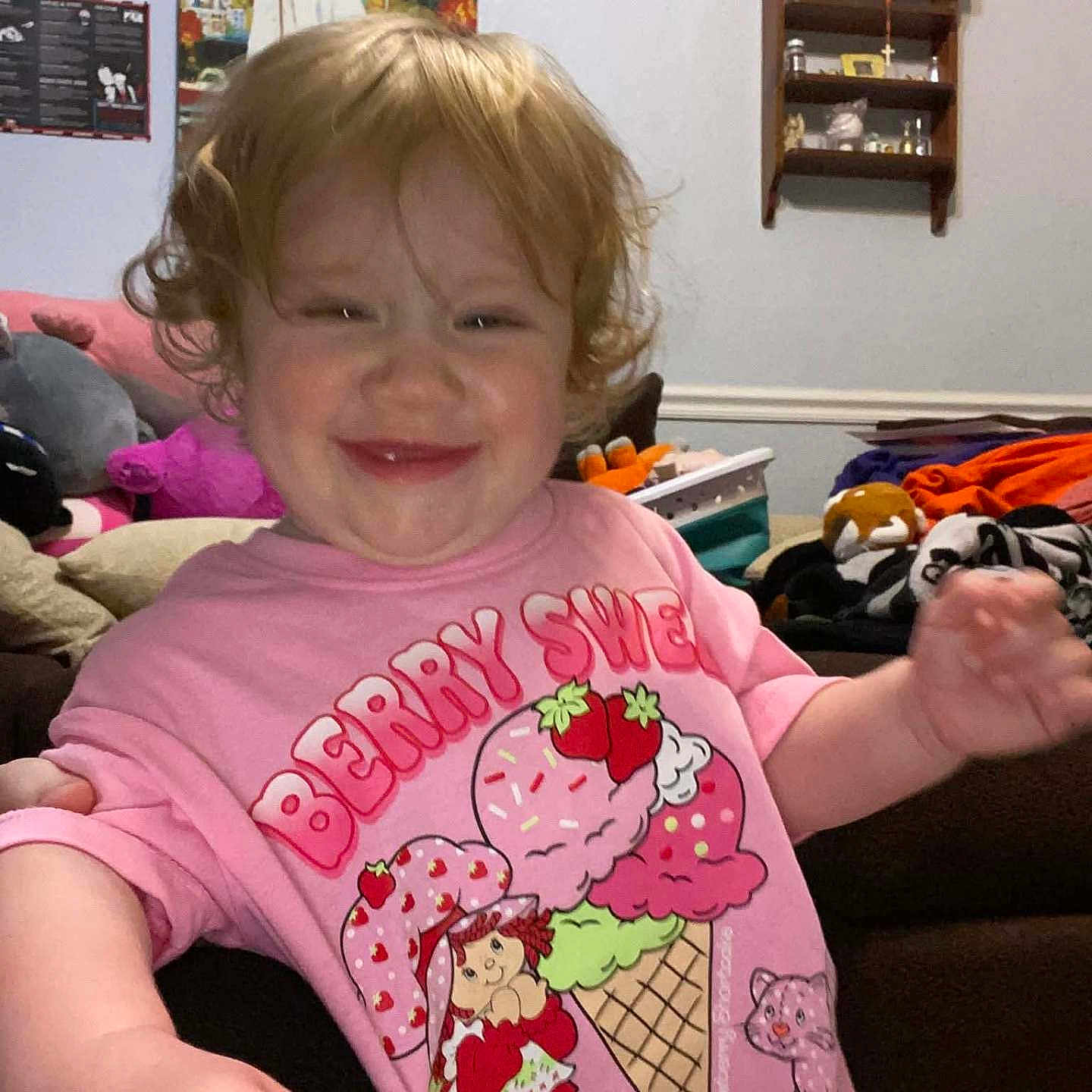 Novaleigh is registered to the contest to win money with this photo: child, toddler, smiling, curly_hair, pink_shirt, graphic_tee, indoor, living_room, toys, couch, wall_shelf, decor, happy, person, face, cute, playful, home, furniture, colorful