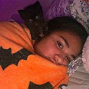 Batman is registered to the contest to win money with this photo: person, dog, blanket, bed, pillow, indoor, portrait, cozy, sleepy, animal, pet, black_dog, orange_blanket, bat_pattern, face, lying_down, comfort, purple_light, snuggle, home