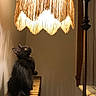 cat, black_cat, radiator, lamp, lampshade, indoor, warm_light, fringe, vintage, furniture, pet, animal, curious, sitting, cozy, shadow, wall, tail, whiskers, looking_up