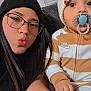 woman, baby, selfie, pacifier, knit_hat, glasses, striped_onesie, couch, indoor, window_blinds, person, child, face, hand, long_hair, black_hat, cute, casual, cozy, smile