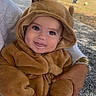baby, child, costume, bear, cute, outdoor, smile, tongue, playful, person, hands, holding, fuzzy, hood, nature, grass, stone_wall, sunlight, casual, daytime