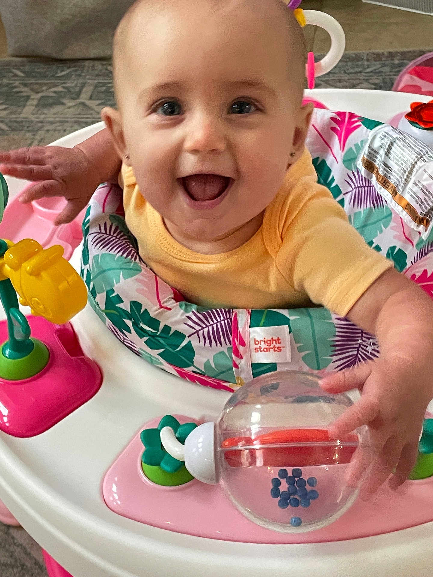 Nora is registered to the contest to win money with this photo: baby, child, smiling, face, eyes, hands, toy, walker, activity_center, colorful, yellow_shirt, happy, indoor, floor, plastic_toy, play, infant, cute, joyful, person