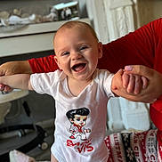 Elvis joined the competition — help win amazing prizes! baby, smile, child, white_onesie, holding_hands, happy, indoor, person, adult_hand, pajamas, christmas_theme, living_room, furniture, television, table, floor, face, cute, infant, joy