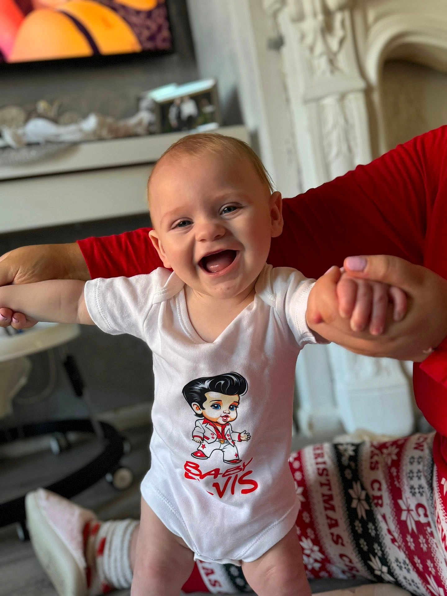 Elvis joined the competition — help win amazing prizes! baby, smile, child, white_onesie, holding_hands, happy, indoor, person, adult_hand, pajamas, christmas_theme, living_room, furniture, television, table, floor, face, cute, infant, joy
