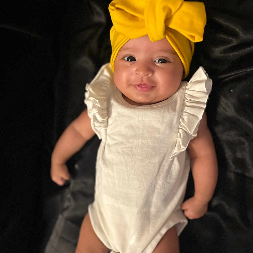 Solana joined the competition — help win amazing prizes! baby, baby_clothes, black_background, child, closeup, cute, expression, face, happy, headgear, indoors, infant, lying_down, one_person, portrait, ruffles, skin, smile, white_outfit, yellow_headwrap