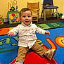 toddler, child, smiling, white_shirt, brown_pants, shoes, playroom, colorful_rug, toys, wooden_chair, plastic_chair, carpet, indoor, happy, cute, person, floor, sitting, baby, play