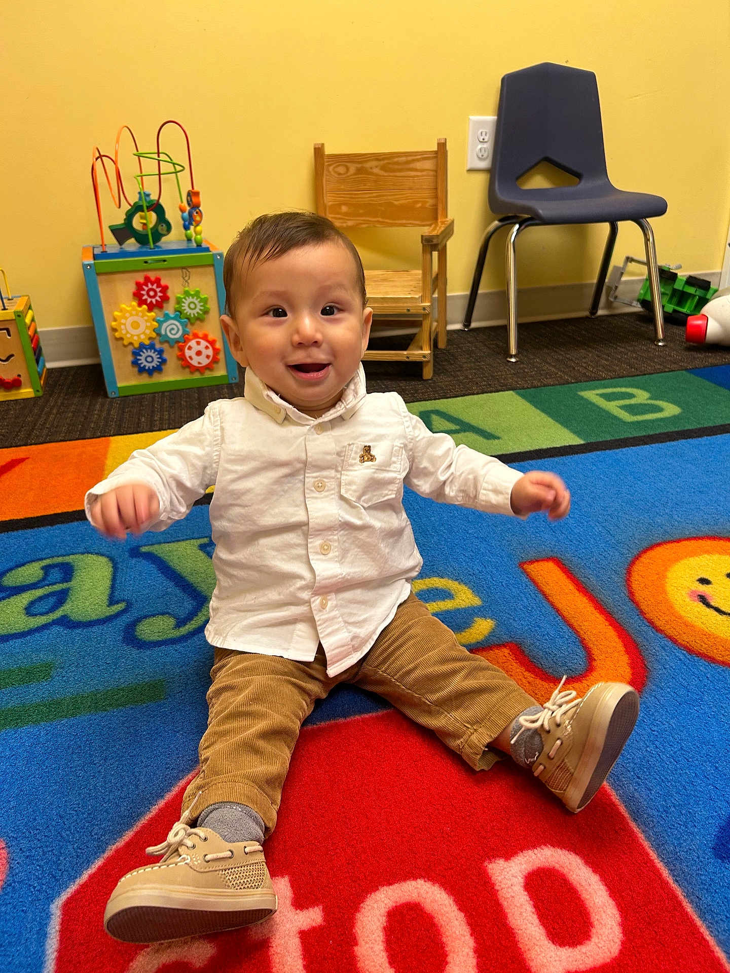 Ismael joined the competition — help win amazing prizes! toddler, child, smiling, white_shirt, brown_pants, shoes, playroom, colorful_rug, toys, wooden_chair, plastic_chair, carpet, indoor, happy, cute, person, floor, sitting, baby, play