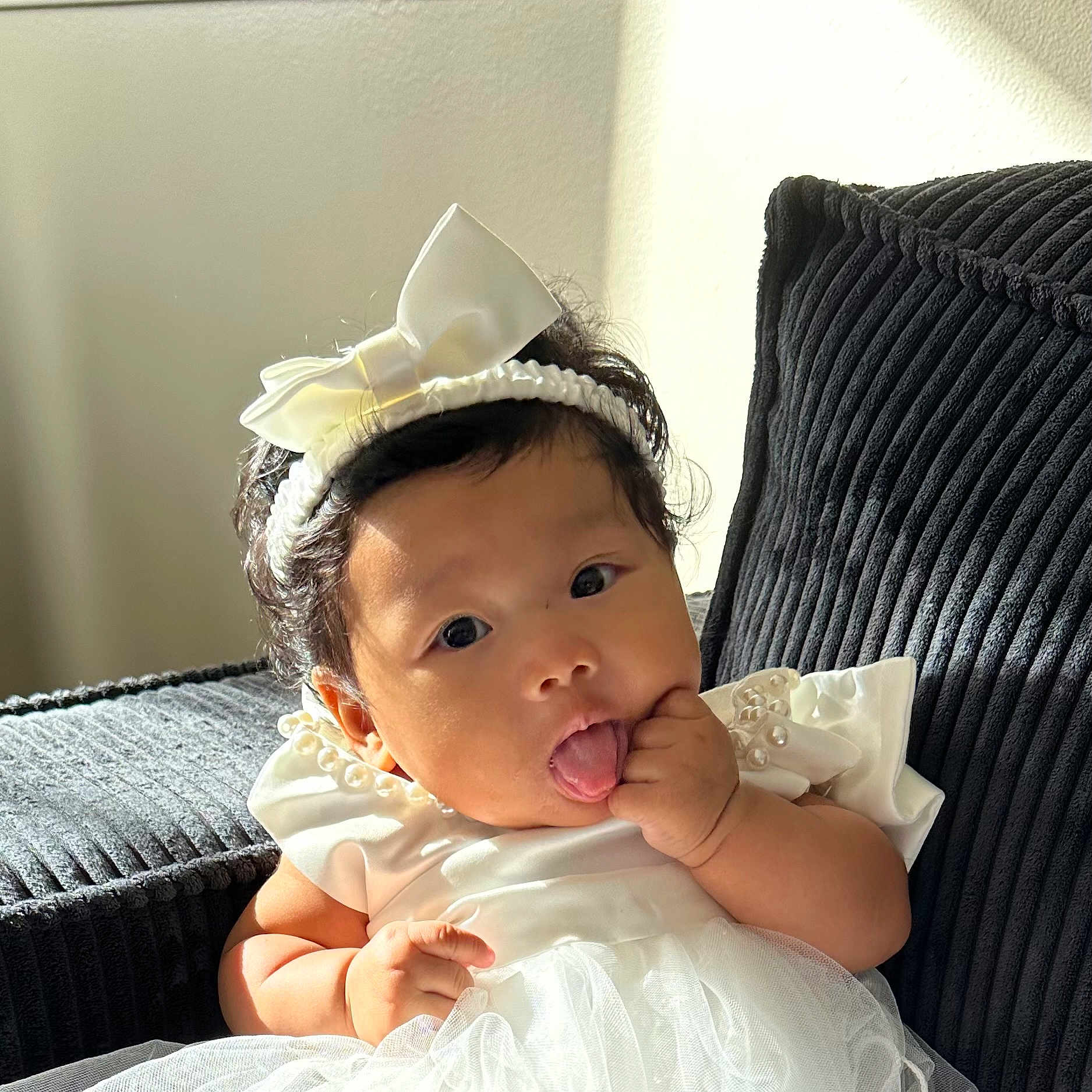 Layana participe au concours pour gagner de l'argent avec cette photo : baby, bow_headband, child, couch, curly_hair, cute, dark_couch, dress, finger_in_mouth, indoor, infant, natural_light, pearl_detail, playful, portrait, sitting, skin, soft_tissue, tongue_out, white_dress
