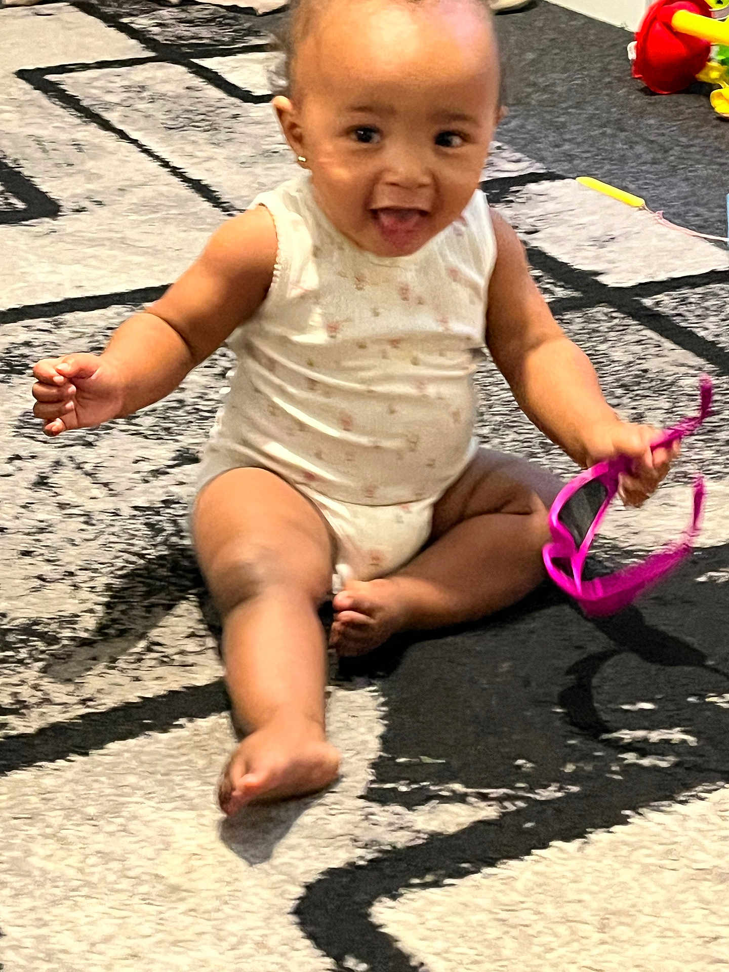Aniyah is registered to the contest to win money with this photo: baby, child, infant, sitting, carpet, floor, pink_sunglasses, toy, happy, smiling, playful, indoor, cute, face, arms, legs, onesie, earrings, tongue, person