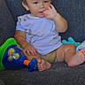 baby, child, toddler, sitting, couch, indoor, portrait, face, smiling, hand, finger, toy, colorful_toy, clothing, shirt, shorts, barefoot, furniture, home, playtime