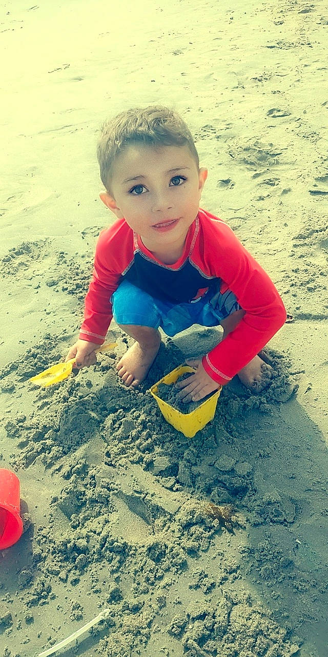 Malakai is registered to the contest to win money with this photo: baby_toddler_clothing, beach, body_of_water, child, electric_blue, face, fun, happy, landscape, leisure, people_in_nature, person, play, recreation, red, sand, sitting, sleeve, soil, t_shirt