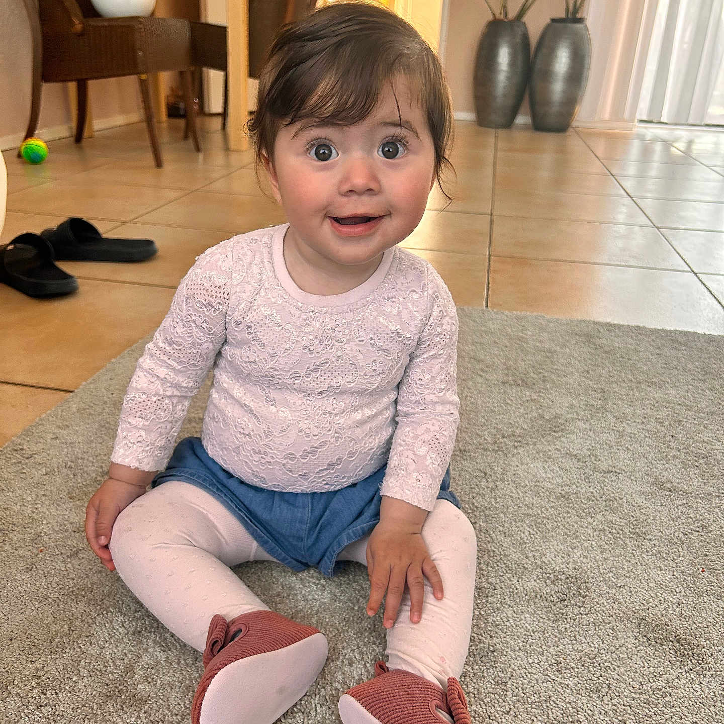 Lara a rejoint le concours — aidez-le/la à gagner de superbes lots ! baby, blue_shorts, booties, carpet, child, cute, expression, face, floor, furniture, home, indoor, light, person, pink_clothing, plant, sitting, surprised, tile_floor, window